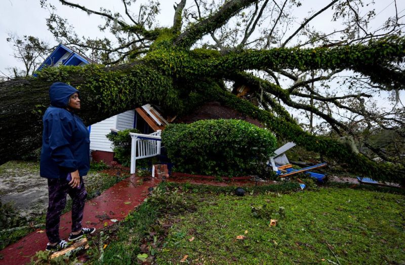 Woman in a rain jacket stands next to tree that has collapsed onto a her home.