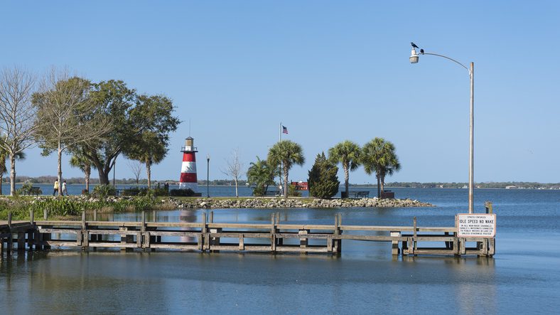 The Mount Dora lighthouse is seen from a nearby road. There are birds on an electrical light pole. An American flag is flying. There are several trees. A wooden dock is in the foreground with a warning sign about no wakes allowed.
